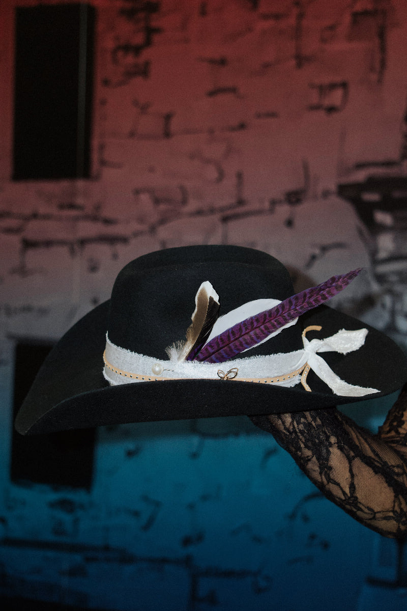 A black hat with white ribbon and brown, purple feathers is held by a lace-gloved hand. Background features a red and blue gradient wall.