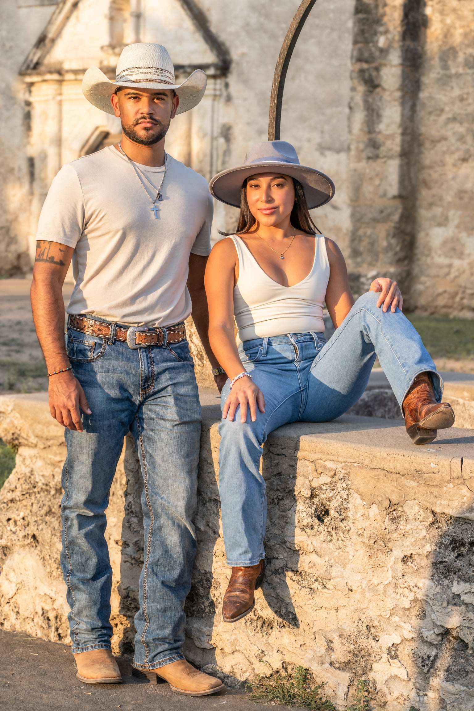 A man and woman in casual Western attire stand against a stone wall. Both wear hats and jeans.