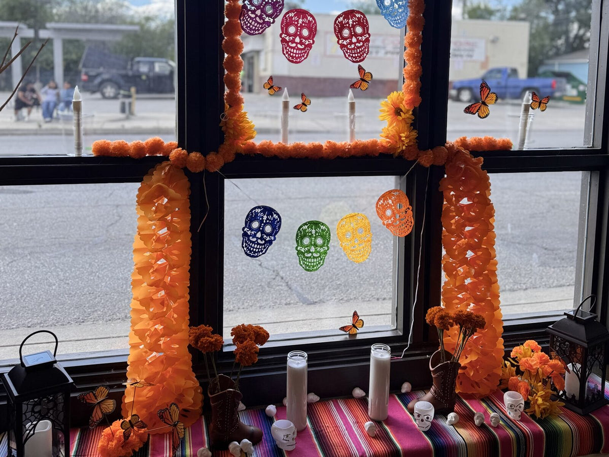 Wild Pony Hat Bar's Day of the Dead ofrenda in a window at Basila Frocks, with marigold garlands, papel picado skulls, candles and cowboy boots