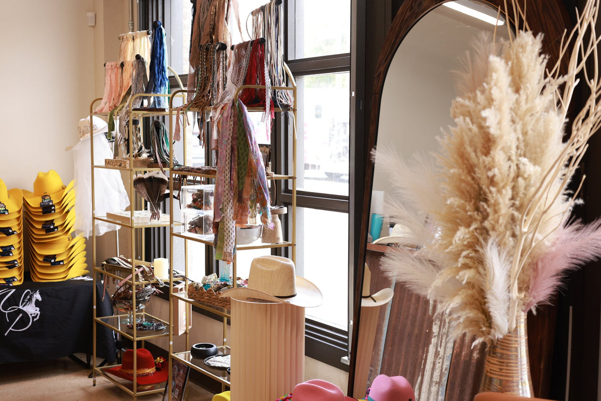 Gold accessories shelf of hat bands beside an arched mirror and pampas grass at Wild Pony Basila Frocks