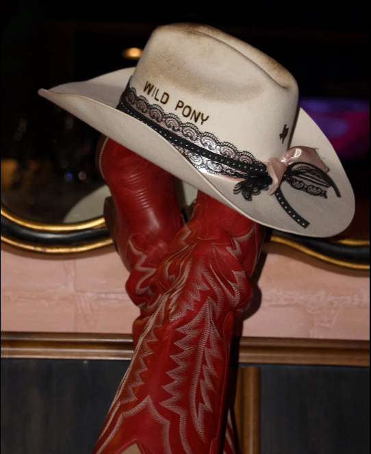 A white cowboy hat labeled "Wild Pony" with black lace and a pink bow rests atop red cowboy boots with intricate stitching, evoking a Western style.