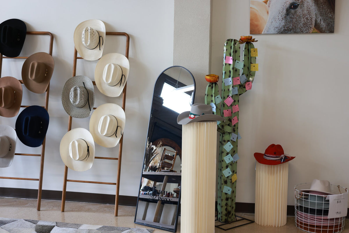 Neutral straw cowboy hats on a ladder rack beside a standing mirror and a cactus note sculpture at Wild Pony Basila Frocks